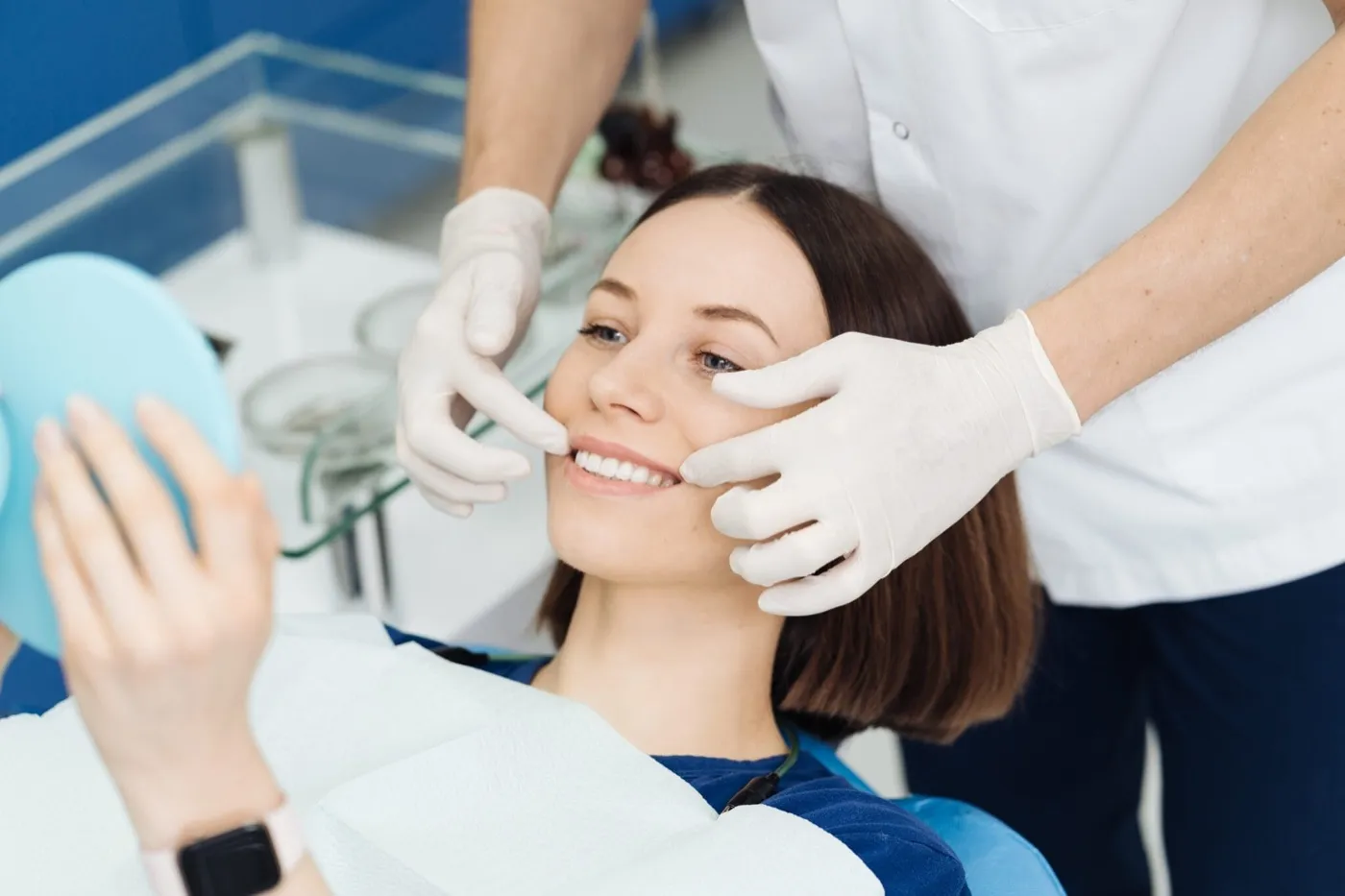A smiling woman in the dental chair holding a hand mirror while a dentist checks her teeth