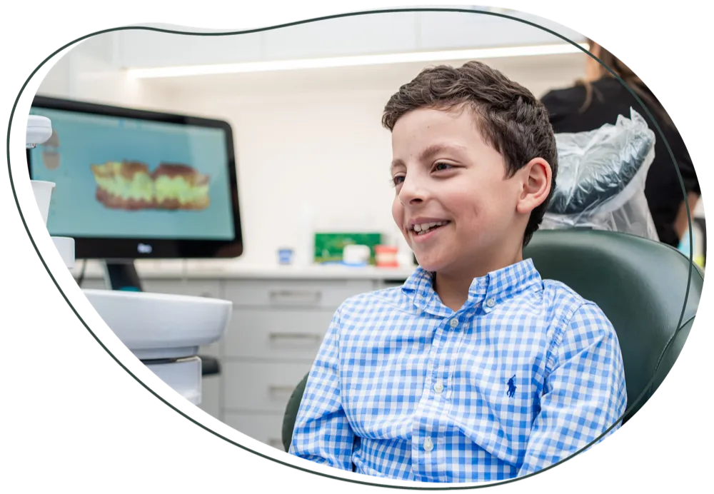 A smiling boy in the dental chair, with a scan of teeth on the screen behind him