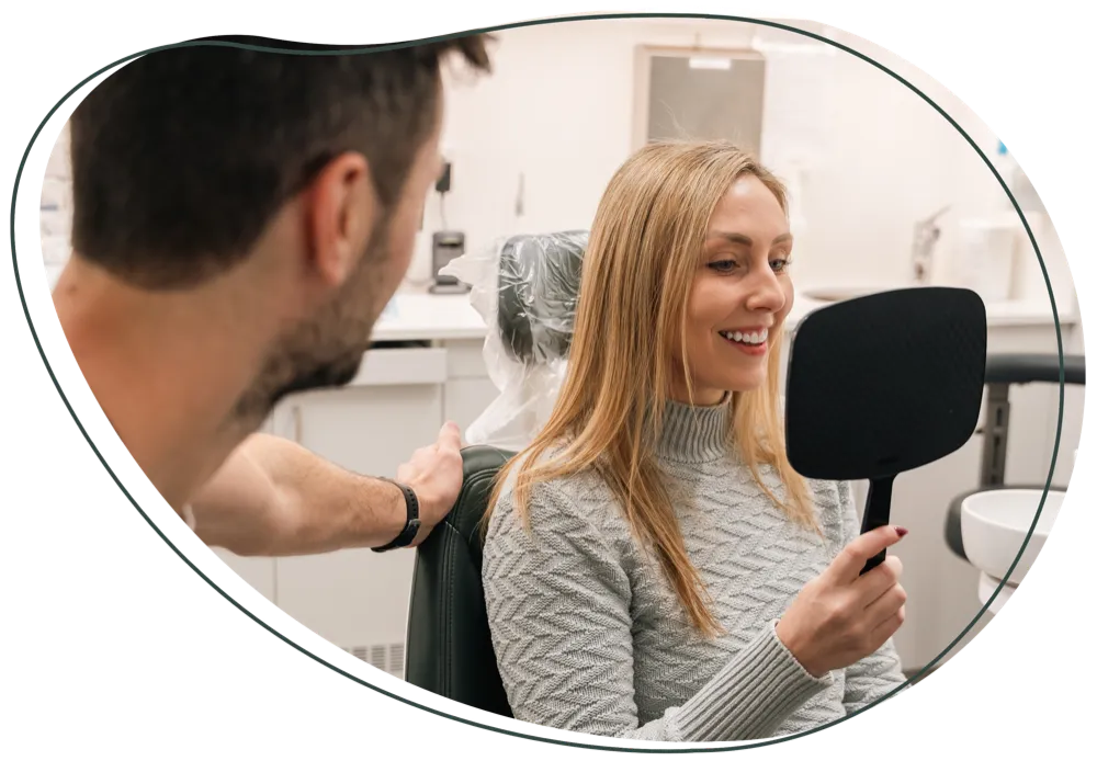 A patient smiling into a hand mirror beside her dentist