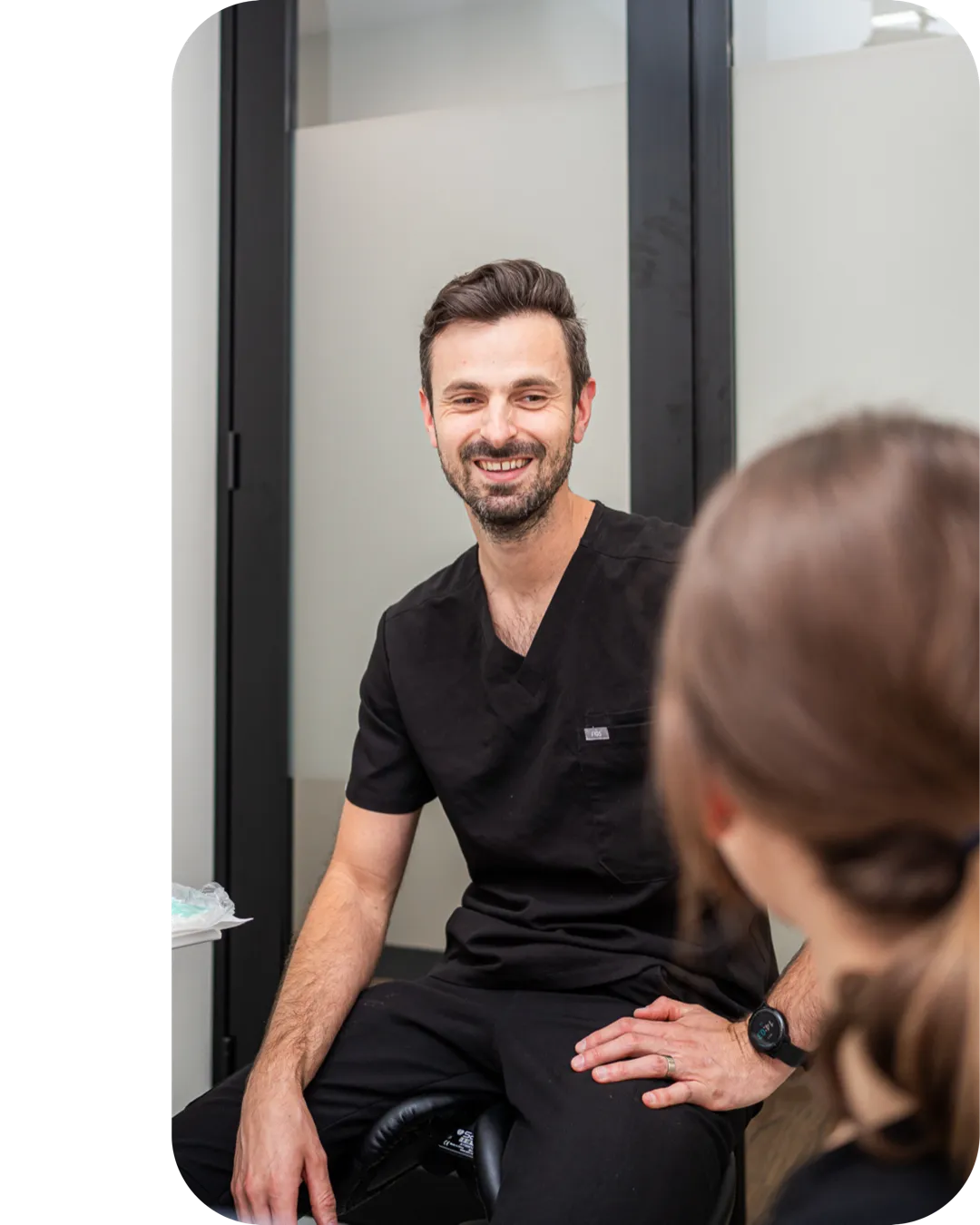 A smiling dentist seated beside a patient in a treatment room